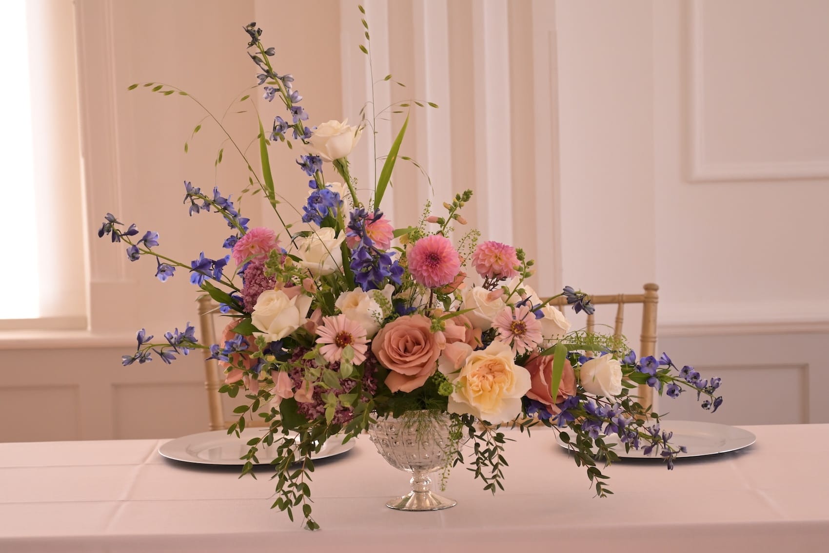 Romantic pink and peach arrangement with roses, blue delphinium and ranunculus on a gold pedestal