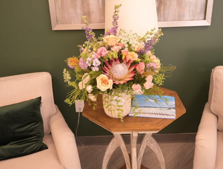 Peach and coral arrangement with king protea, garden roses and purple larkspur on a wooden side table