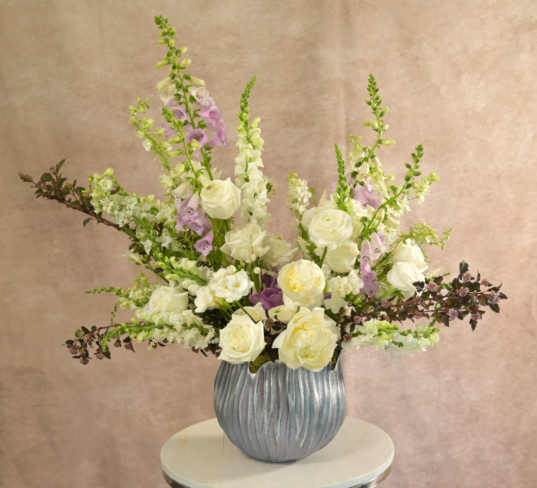 White roses with lavender stock and green viburnum accented by purple florals in a clear glass vase